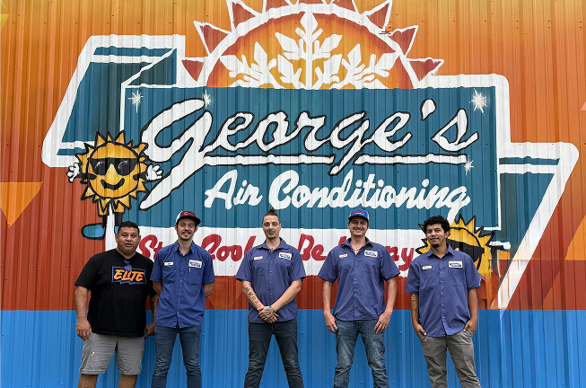 Five men stand in front of a colorful "George’s Air Conditioning" sign painted on a corrugated metal wall, with four wearing matching work shirts and one in a black t-shirt.